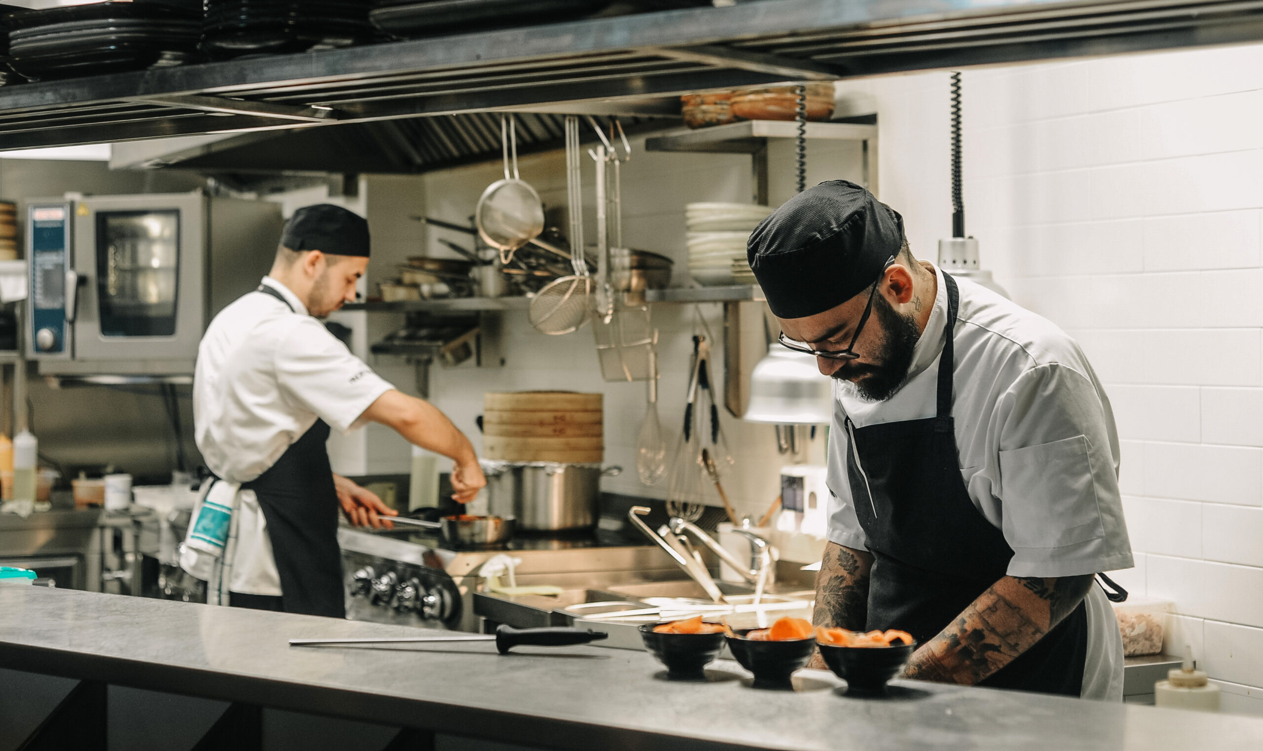 En restaurangmiljö från köket där det står två kockar och tillreder mat